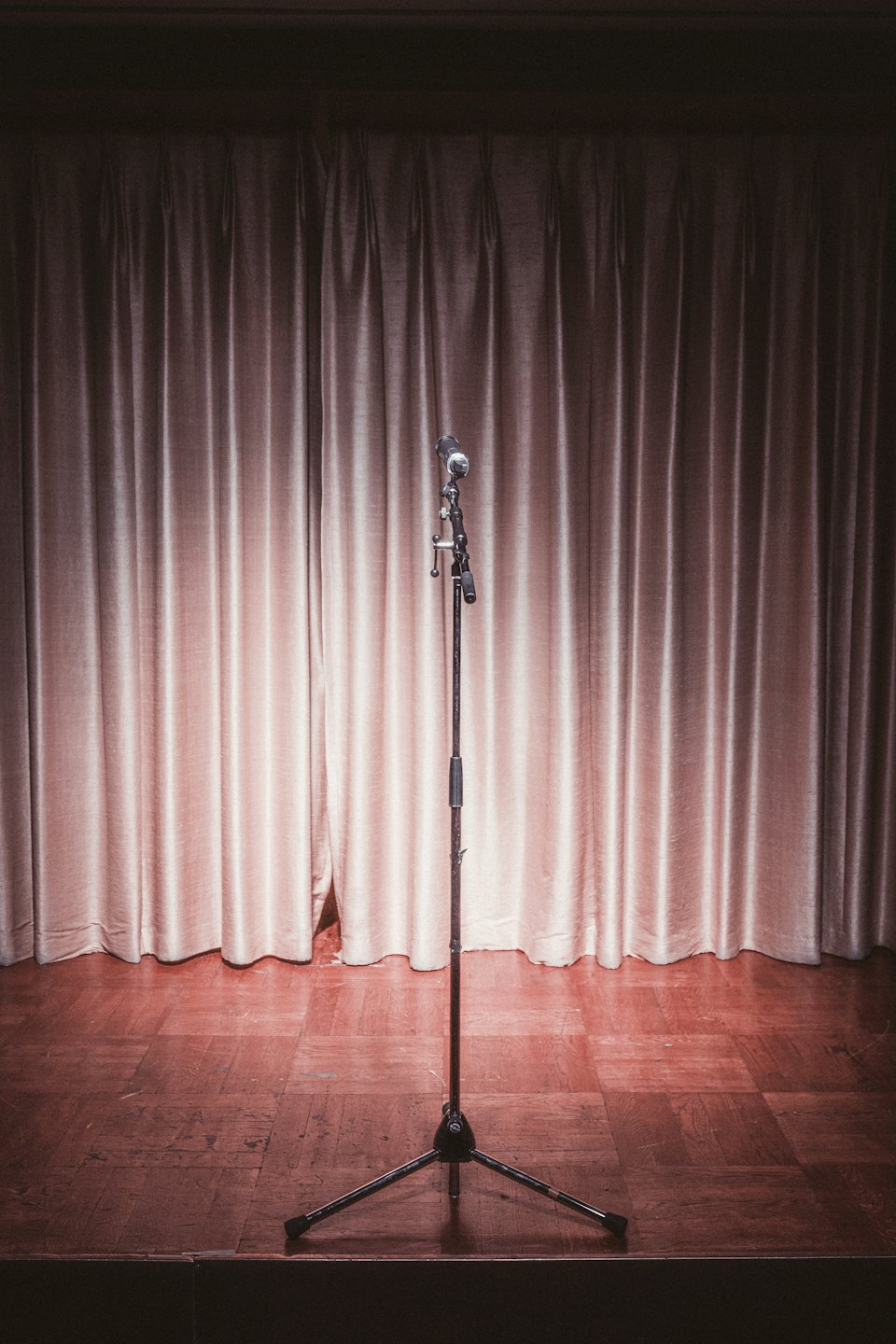 Un supporto per microfono al centro di un palco di legno davanti a tende beige.